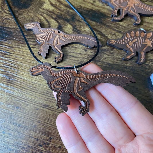 A wooden Utahraptor skeleton necklace held in a person's hand, with additional matching wooden dinosaur skeleton pendants on a black cord in the background.